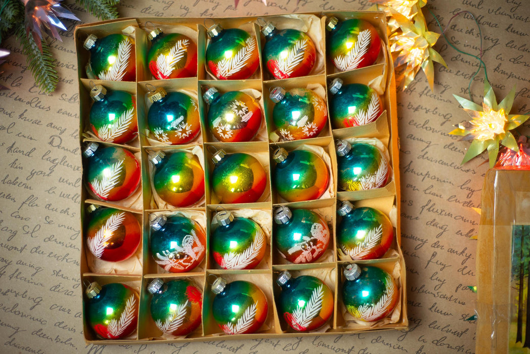 A box filled with colorful Christmas ornaments, including glass baubles and star-shaped ornaments, placed on a surface with some holiday decorations and a piece of paper with handwriting.