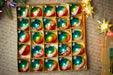 A box filled with colorful Christmas ornaments, including glass baubles and star-shaped ornaments, placed on a surface with some holiday decorations and a piece of paper with handwriting.