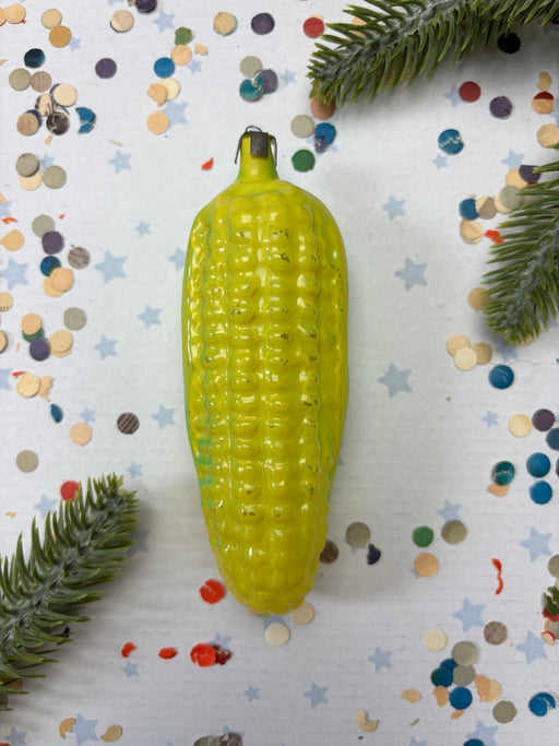 A yellow corn cob ornament is placed on a white background with scattered confetti and pine branches.