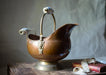 A brass-colored metal pitcher with a decorative handle sits on a wooden surface, accompanied by a red book and a white ribbon.