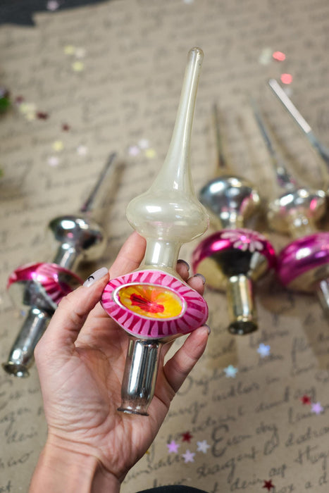 A hand holding a decorative ornament with a pink and yellow design, surrounded by other ornaments in the background.