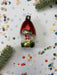 A red ornament with a green base is placed on a white background, surrounded by scattered confetti and a sprig of greenery.