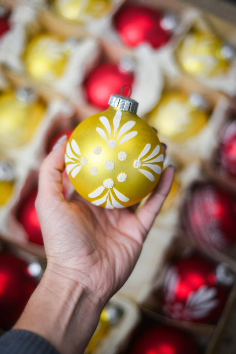 A hand holding a yellow ornament with white floral designs against a background of red and yellow ornaments.