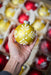 A hand holding a yellow ornament with white floral designs against a background of red and yellow ornaments.