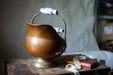 An old, copper-colored metal bucket with a blue and white floral handle sits on a wooden surface, accompanied by a red book and a white ribbon.