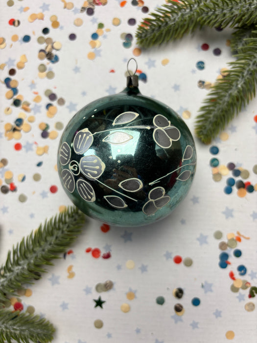 A black ornament with white floral designs is surrounded by colorful confetti and a sprig of greenery on a white background.