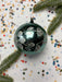 A black ornament with white floral designs is surrounded by colorful confetti and a sprig of greenery on a white background.
