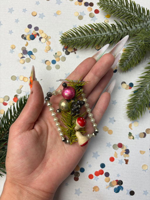 A hand holding a decorative bracelet made of beads and pine branches, surrounded by confetti and pine branches.
