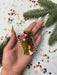 A hand holding a decorative bracelet made of beads and pine branches, surrounded by confetti and pine branches.