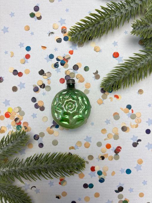 a green ornament with a star design, surrounded by colorful confetti and pine branches.