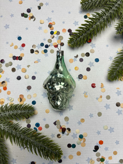 a green glass ornament with a twisted shape, surrounded by colorful confetti and pine branches.
