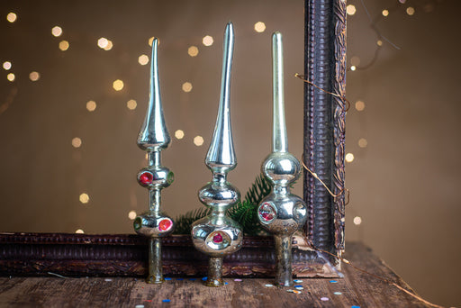 Three ornate Christmas tree ornaments with red and silver accents are displayed on a wooden surface, with a blurred background of twinkling lights.