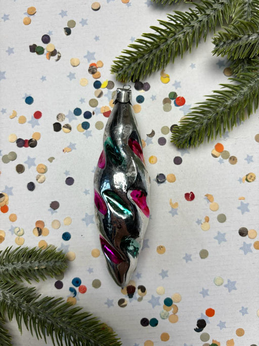A shiny, metallic ornament with a twisted shape and colorful accents is surrounded by confetti and pine branches on a white background.