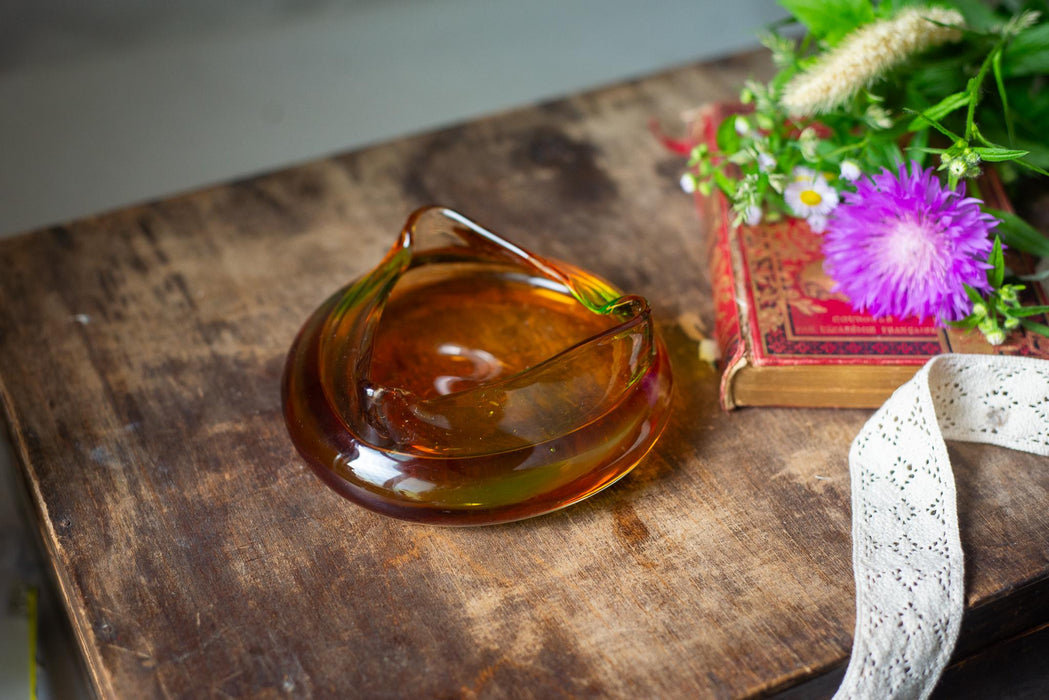 A glass ashtray sits on a wooden surface, with a bouquet of flowers and a book nearby.