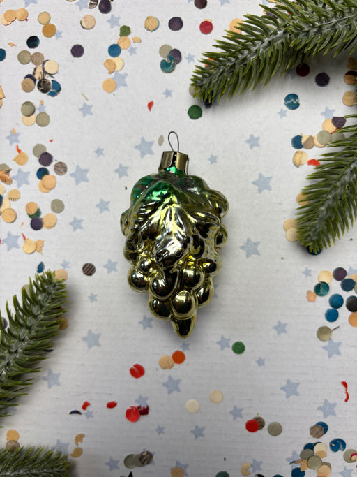 A shiny green ornament resembling a bunch of grapes is placed on a white background with scattered confetti and pine branches.