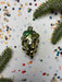 A shiny green ornament resembling a bunch of grapes is placed on a white background with scattered confetti and pine branches.