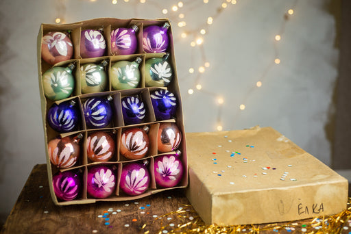 A cardboard box filled with colorful Christmas ornaments, including purple, green, blue, and orange balls, along with confetti scattered around the box.