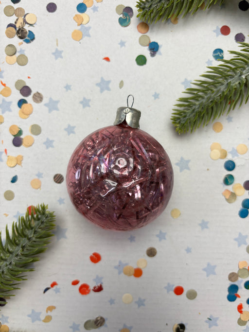 A pink glass ornament with a silver clasp is surrounded by confetti and a sprig of greenery on a white background.