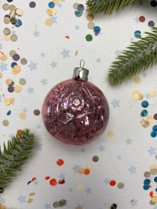 A pink glass ornament with a silver clasp is surrounded by confetti and a sprig of greenery on a white background.