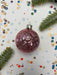 A pink glass ornament with a silver clasp is surrounded by confetti and a sprig of greenery on a white background.