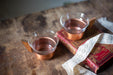 Two copper-colored glasses filled with a clear liquid sit on a wooden surface, accompanied by a red book with a lace-like cover.