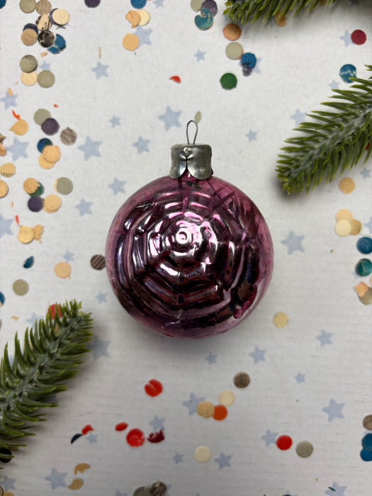 A purple glass ornament with a silver clasp is surrounded by confetti and a sprig of greenery on a white background.