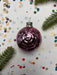 A purple glass ornament with a silver clasp is surrounded by confetti and a sprig of greenery on a white background.