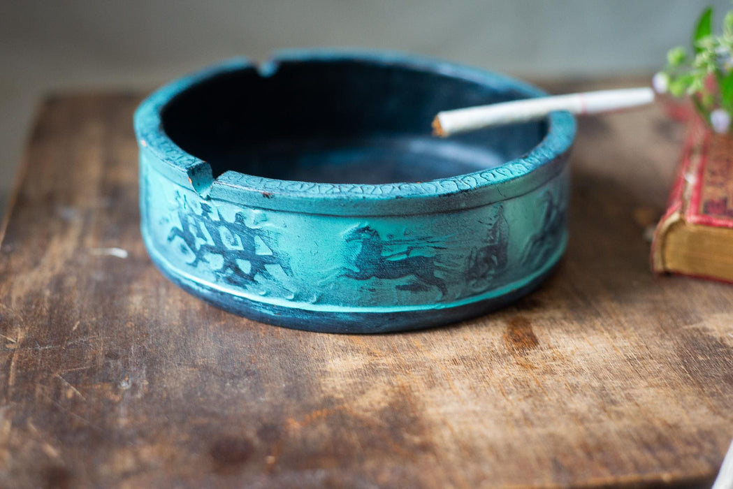 A blue ashtray with a decorative design sits on a wooden surface next to a lit cigarette.