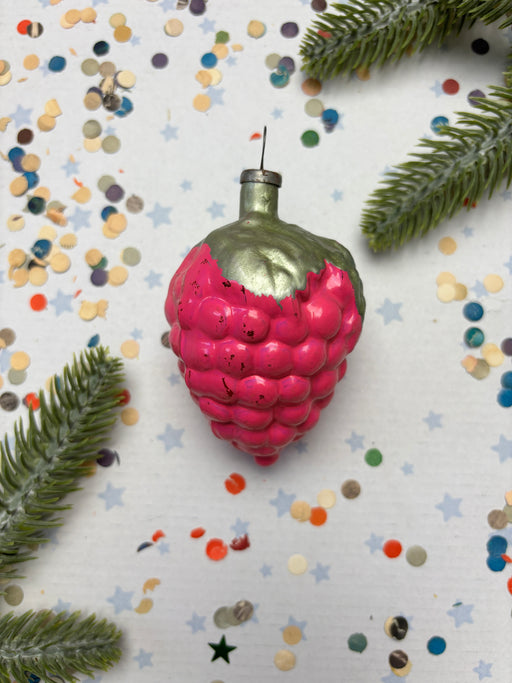 A pink, raspbery ornament with a green base is surrounded by colorful confetti and pine branches on a white background.