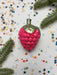 A pink, raspbery ornament with a green base is surrounded by colorful confetti and pine branches on a white background.