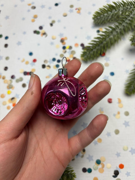 A hand holding a small, shiny pink ornament against a background of colorful confetti and pine branches.