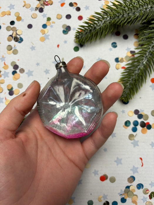 A hand holding a clear glass ornament with a pink and purple design, surrounded by colorful confetti on a white background.
