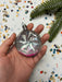 A hand holding a clear glass ornament with a pink and purple design, surrounded by colorful confetti on a white background.