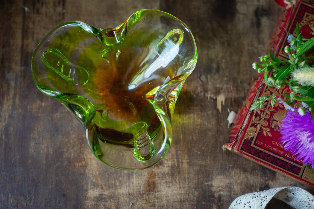 a green glass vase with a flower arrangement on a wooden surface, accompanied by a book.
