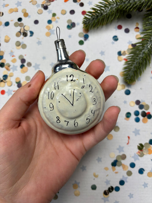 A hand holding a small white clock ornament with black numbers and hands, set against a background of colorful confetti.