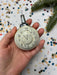 A hand holding a small white clock ornament with black numbers and hands, set against a background of colorful confetti.
