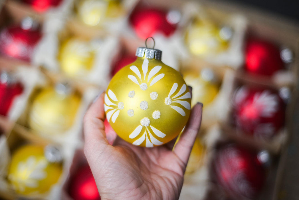 A hand holding a yellow ornament with white floral designs against a background of red and yellow ornaments.