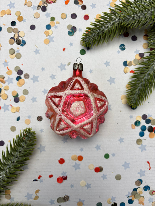 A red star-shaped ornament with a silver star at the center is surrounded by pine branches, confetti, and small star-shaped decorations on a white background.