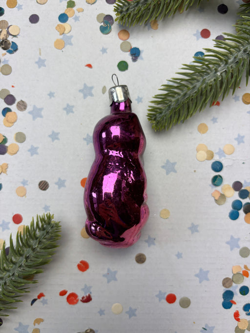 A purple glass ornament is placed on a white background, surrounded by confetti and green pine branches.