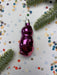 A purple glass ornament is placed on a white background, surrounded by confetti and green pine branches.