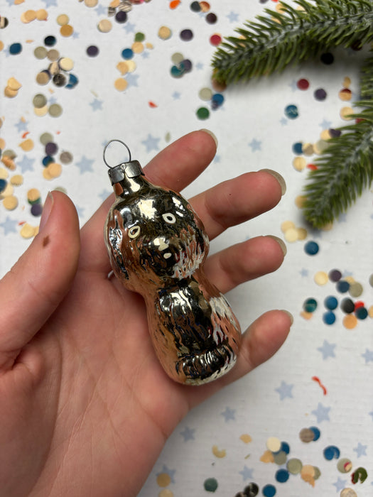 A hand holding a shiny, metallic ornament resembling a bear or a similar animal shape, with a background of colorful confetti or small circles.