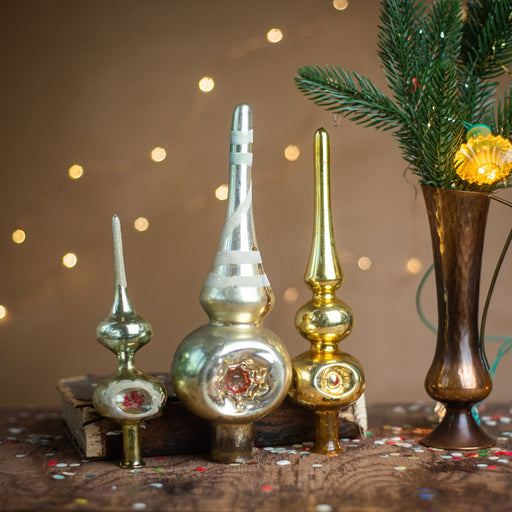 a collection of vintage ornaments, including a tall, cylindrical ornament with a pointed top, a smaller, round ornament, and a tall, conical ornament, all displayed on a wooden surface. A vase with a green plant and twinkling lights can be seen in the background, creating a festive atmosphere.