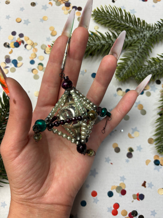 A hand holding a beaded necklace with a star-shaped pendant, surrounded by confetti and a pine tree branch.