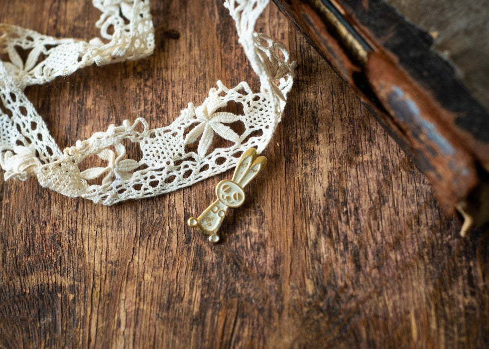 A delicate white lace trim and a small golden rabbit charm are placed on a wooden surface.
