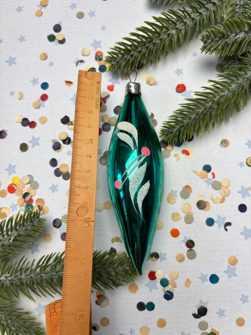 A green glass ornament with a white design is placed next to a ruler on a background of confetti and pine branches.