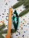 A green glass ornament with a white design is placed next to a ruler on a background of confetti and pine branches.