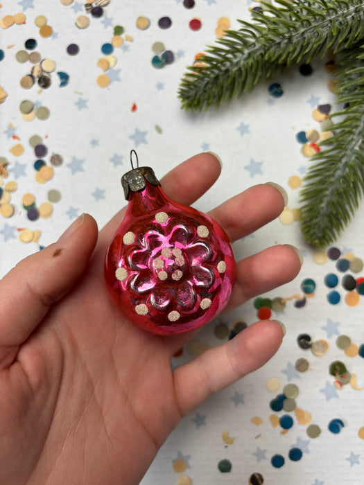A hand holding a pink ornament with a flower design, surrounded by confetti and a pine branch.