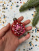 A hand holding a pink ornament with a flower design, surrounded by confetti and a pine branch.