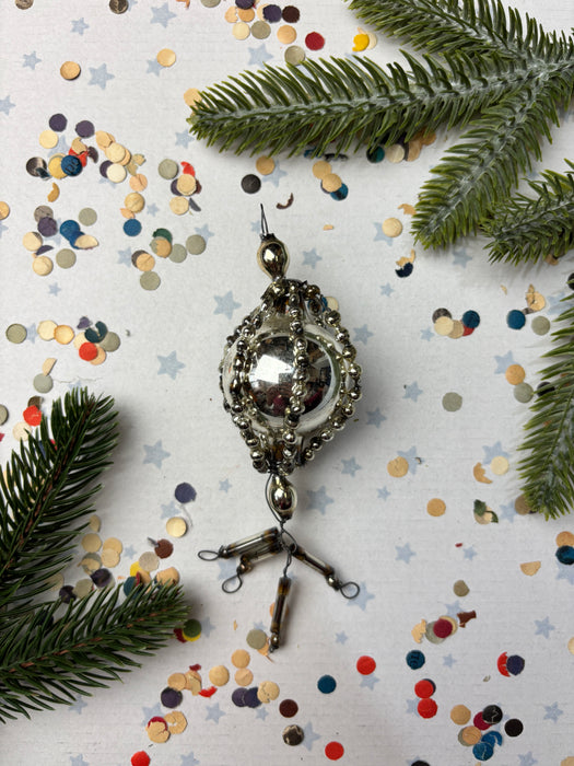 a silver ornament with a round shape and a metallic design, surrounded by pine branches, confetti, and star-shaped decorations on a white background.