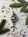 a silver ornament with a round shape and a metallic design, surrounded by pine branches, confetti, and star-shaped decorations on a white background.